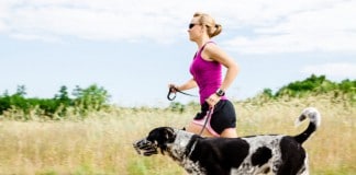 Sommerfitness mit Hund: Eine Läuferin mit pinkfarbenem Top läuft mir ihrem Hund an einem Feld mit hohem trocken-gelben Gras vorbei.