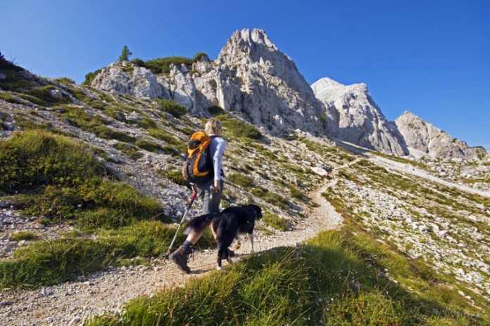 Eine junge Frau geht mit ihrem Hund einen Bergpfad aufwärts, steinige Gipfel im Hintergrund, rechts Abhang mit Gras.